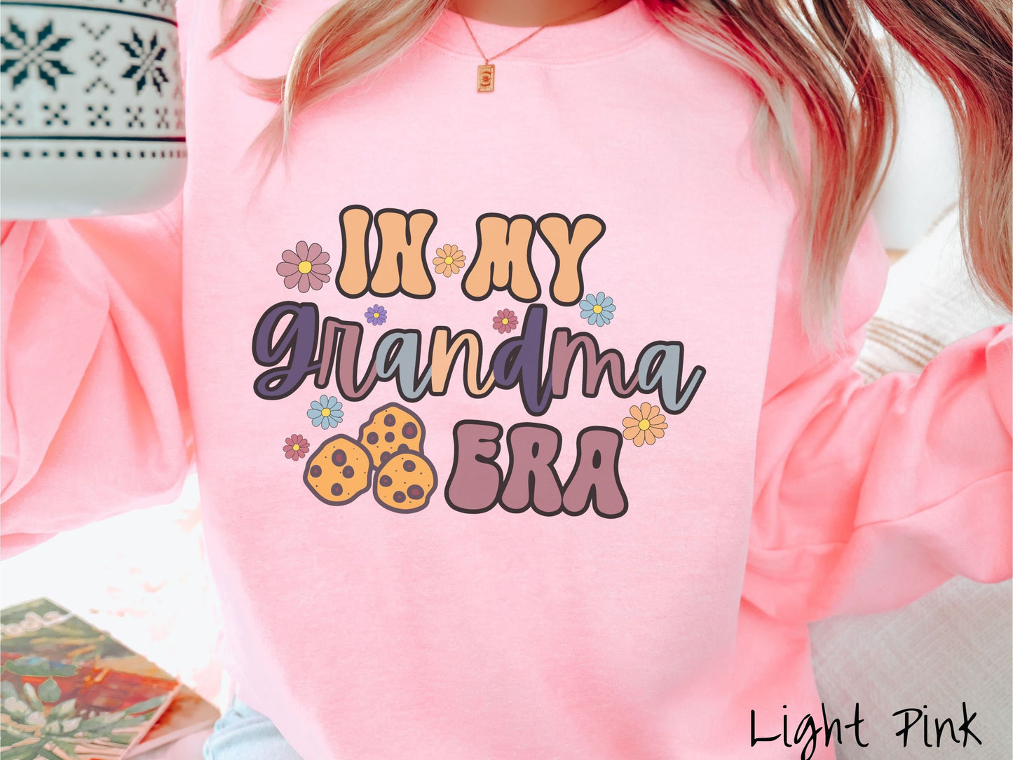 A woman wearing a cute light pink colored sweatshirt with text on the front saying In My Grandma Era, with flowers and chocolate chip cookies mixed within the text.