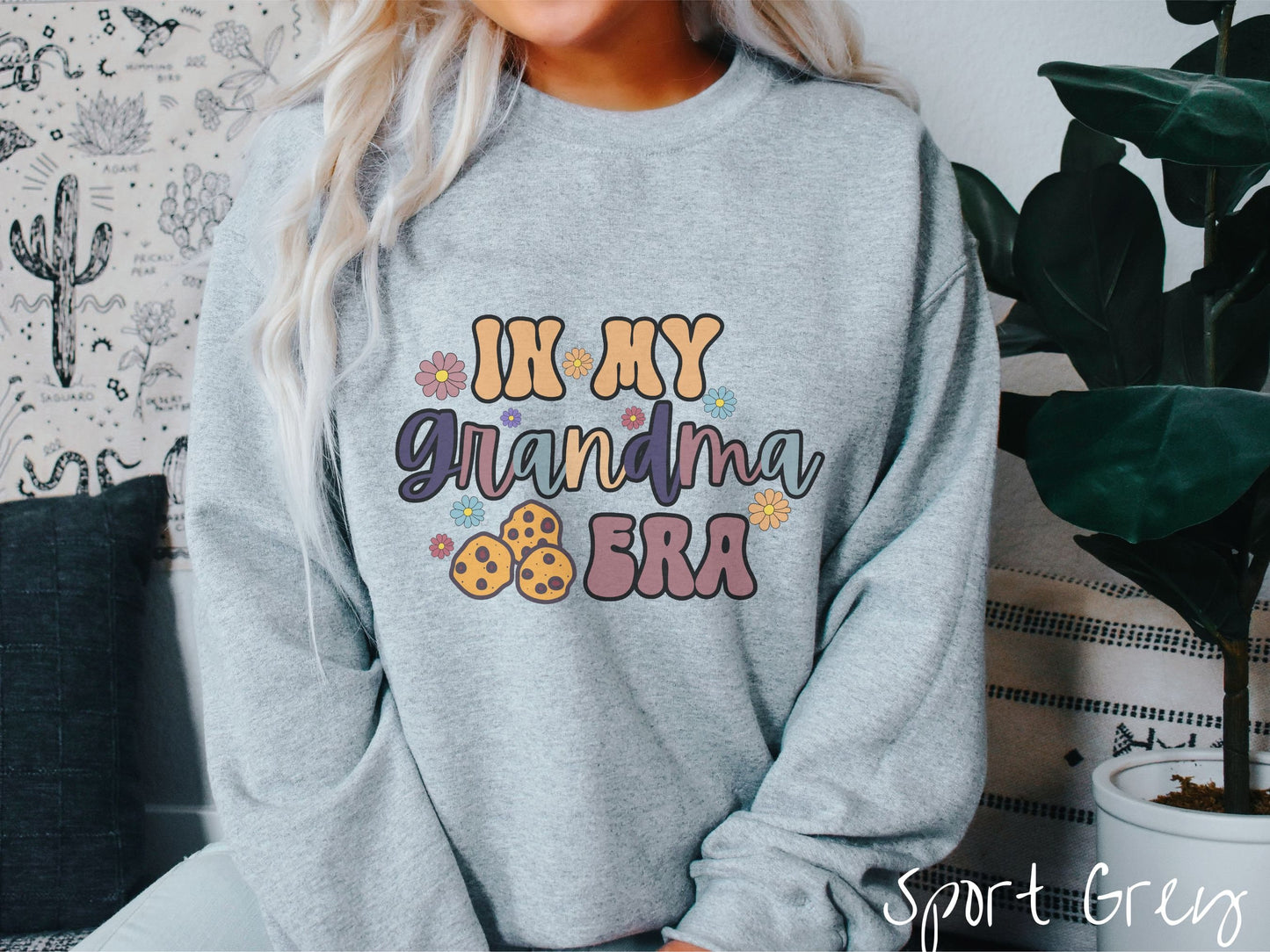 A woman wearing a cute sport grey colored sweatshirt with text on the front saying In My Grandma Era, with flowers and chocolate chip cookies mixed within the text.
