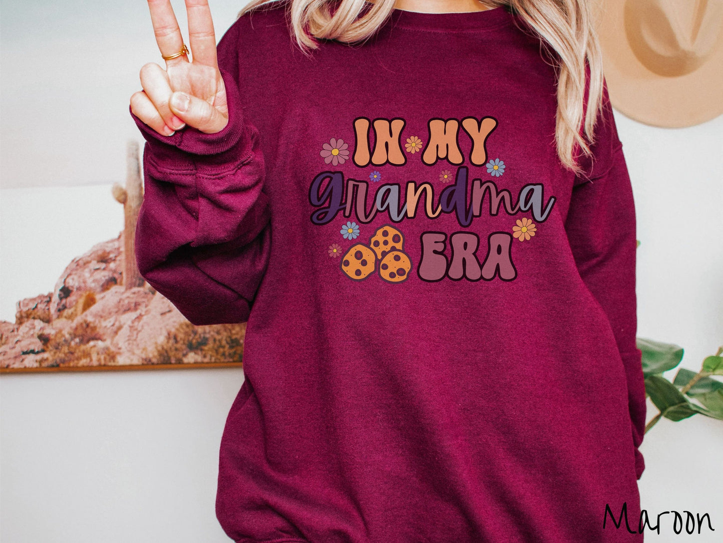 A woman wearing a cute maroon colored sweatshirt with text on the front saying In My Grandma Era, with flowers and chocolate chip cookies mixed within the text.