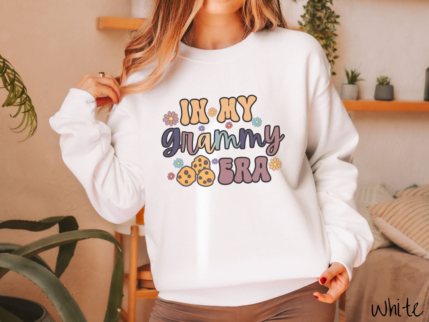 A woman wearing a cute white sweatshirt with the text saying In My Grammy era with flowers and chocolate chip cookies mixed within the text.