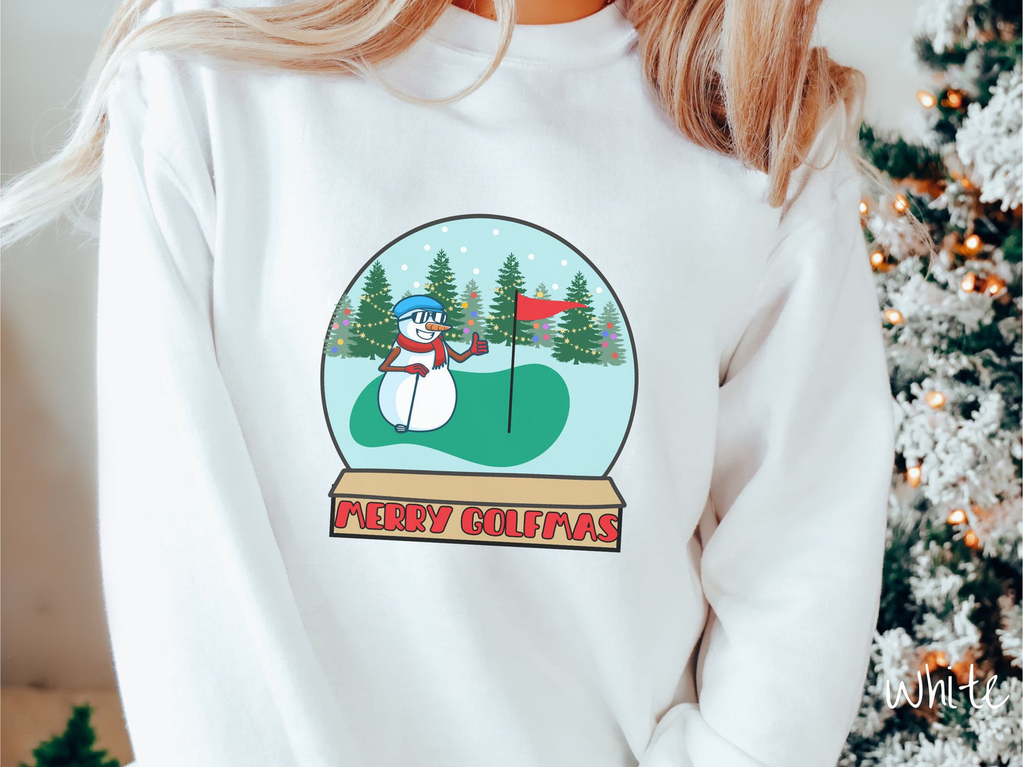 A woman wearing a cute white colored sweatshirt with red text saying Merry Golfmas and a picture of a snowman giving the thumbs up on the golf green next to the hole with a flag in it.