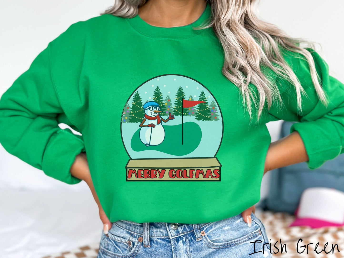A woman wearing a cute Irish green colored sweatshirt with red text saying Merry Golfmas and a picture of a snowman giving the thumbs up on the golf green next to the hole with a flag in it.
