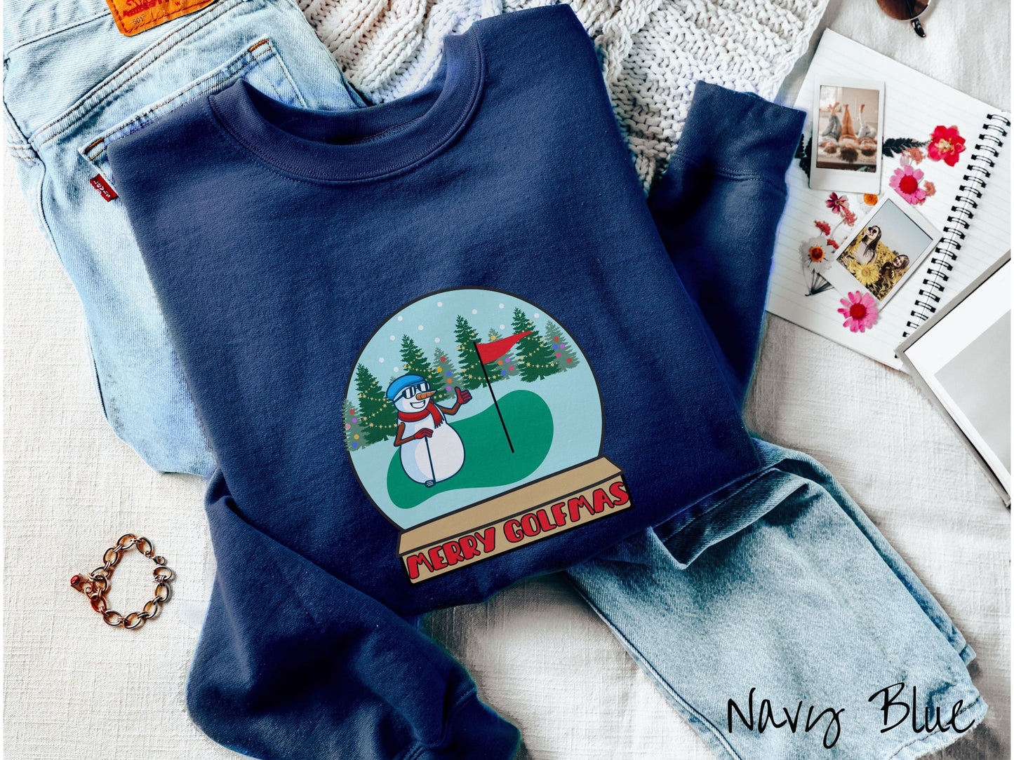 A cute navy blue colored sweatshirt with red text saying Merry Golfmas and a picture of a snowman giving the thumbs up on the golf green next to the hole with a flag in it.