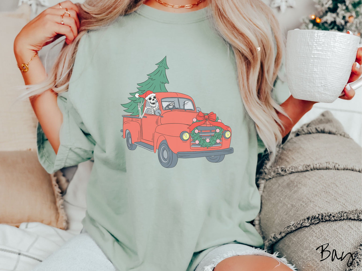 A woman wearing a cute, vintage bay colored comfy sweatshirt with a skeleton wearing a Christmas santa hat driving an old red pickup truck with a Christmas tree stuffed into the back, and a wreath across the front bumper.