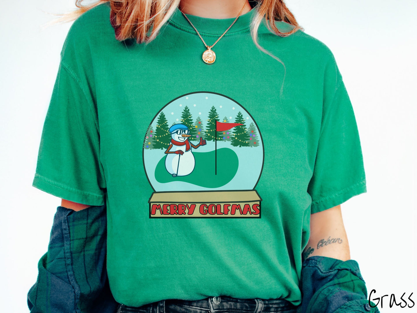A woman wearing a cute green colored shirt with red text saying Merry Golfmas and a picture of a snowman giving the thumbs up on the golf green next to the hole with a flag in it.