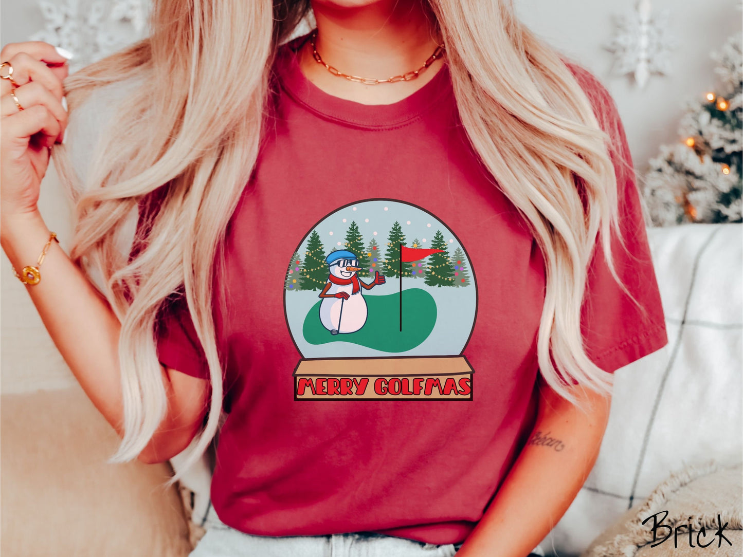 A woman wearing a cute brick colored shirt with red text saying Merry Golfmas and a picture of a snowman giving the thumbs up on the golf green next to the hole with a flag in it.