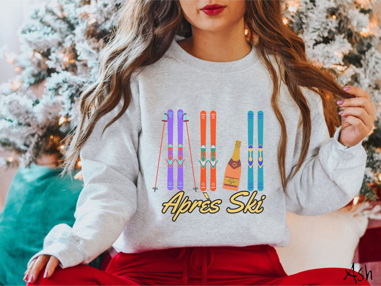 A woman wearing a vintage, ash colored sweatshirt with sets of purple, orange, and turquoise snow skis standing upright, and a big bottle of champagne next to them. Below the skis is the text Apres Ski which is French for After Skiing.