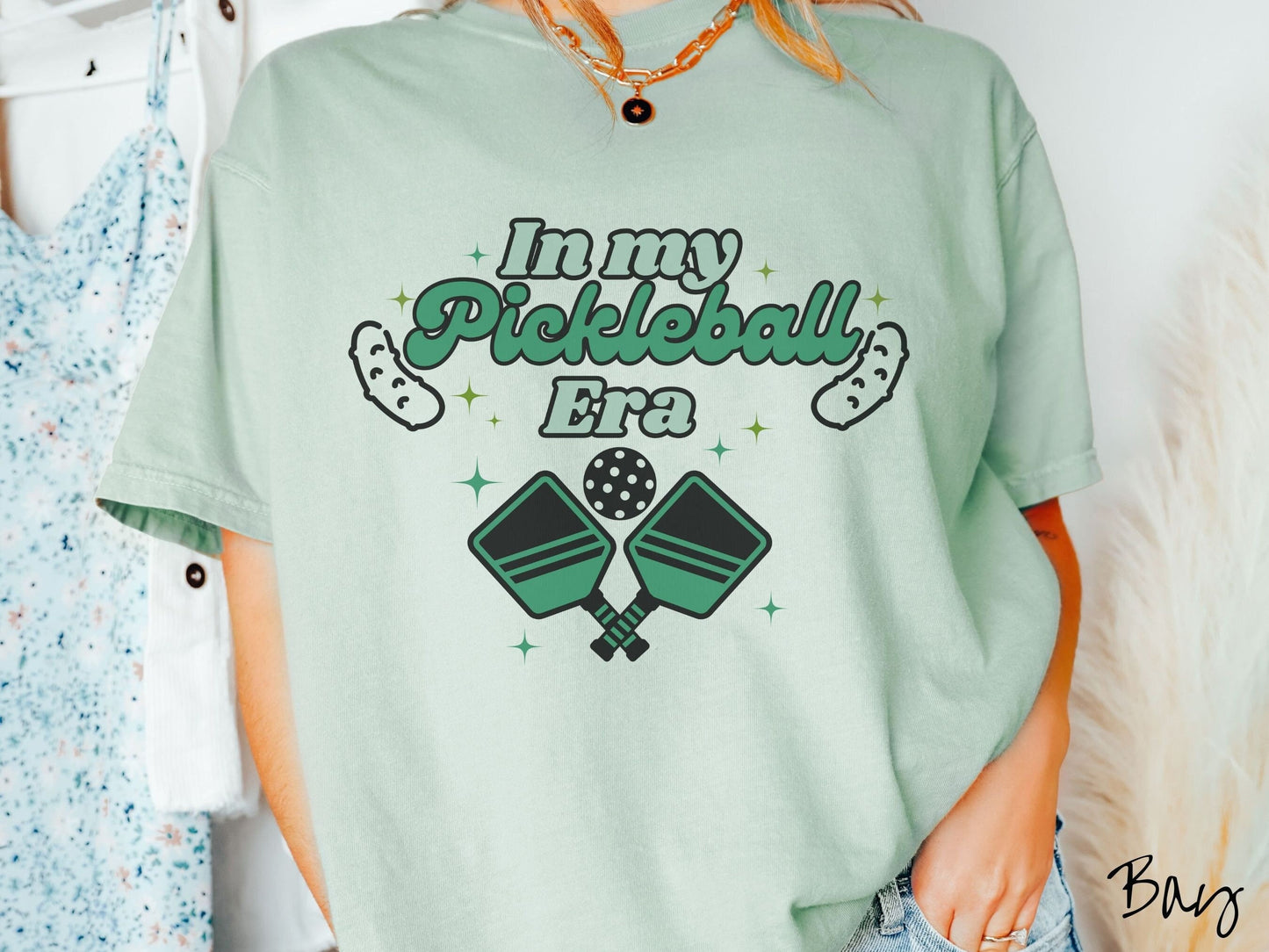 A woman wearing a cute bay colored shirt with the text In My Pickleball Era across the top, and below are 2 pickleball paddles and a ball. There are pickles at the bottom left and right corners of the text.