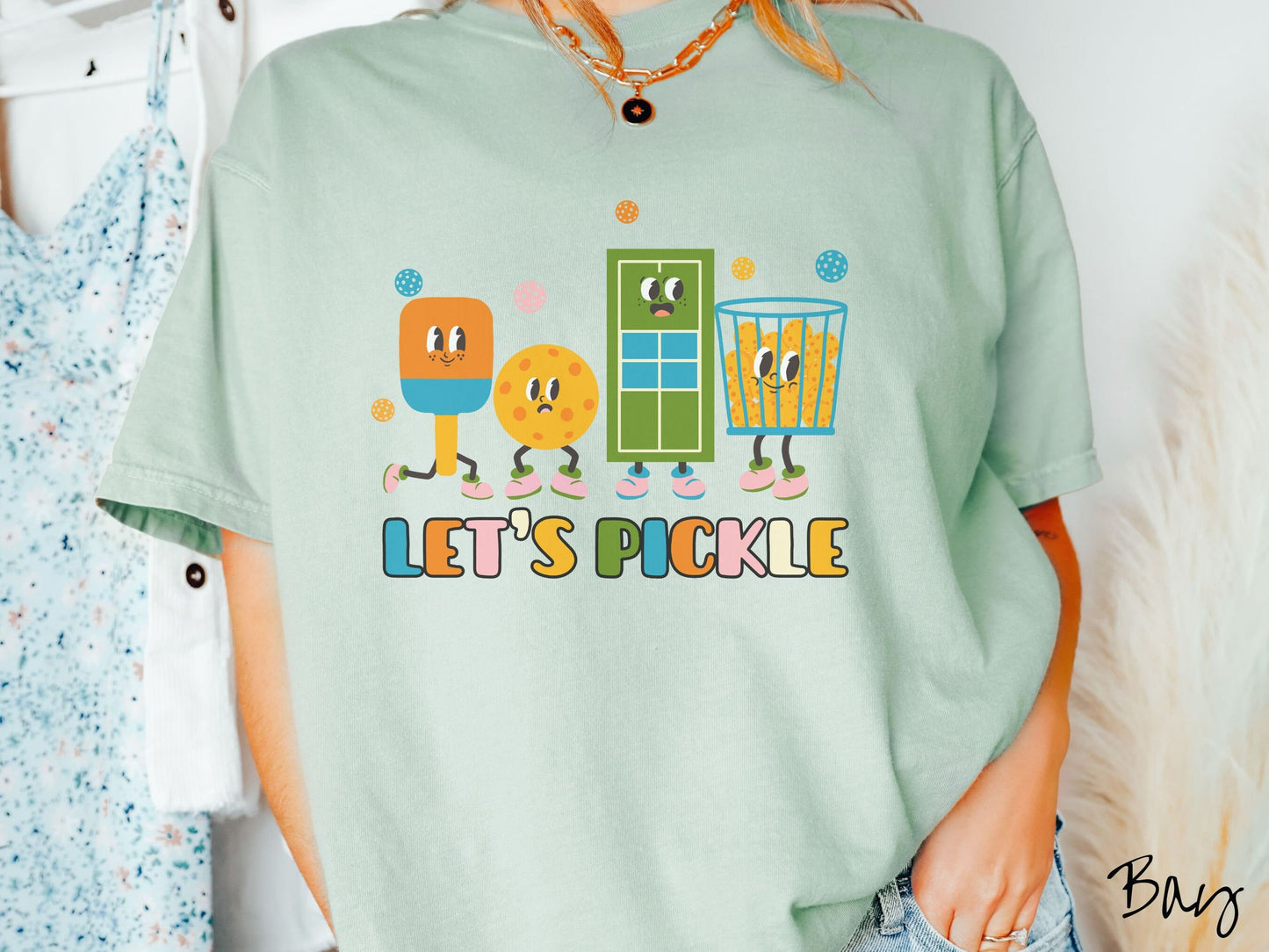 A woman wearing a cute bay colored shirt with the text Let's Pickle along the bottom, and above the text are a popsicle, cookie, popcorn, and pickle balls.
