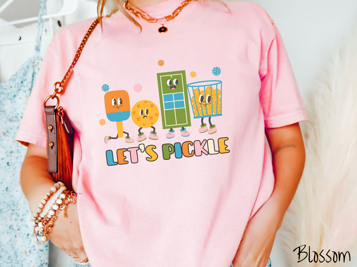 A woman wearing a cute blossom colored shirt with the text Let's Pickle along the bottom, and above the text are a popsicle, cookie, popcorn, and pickle balls.