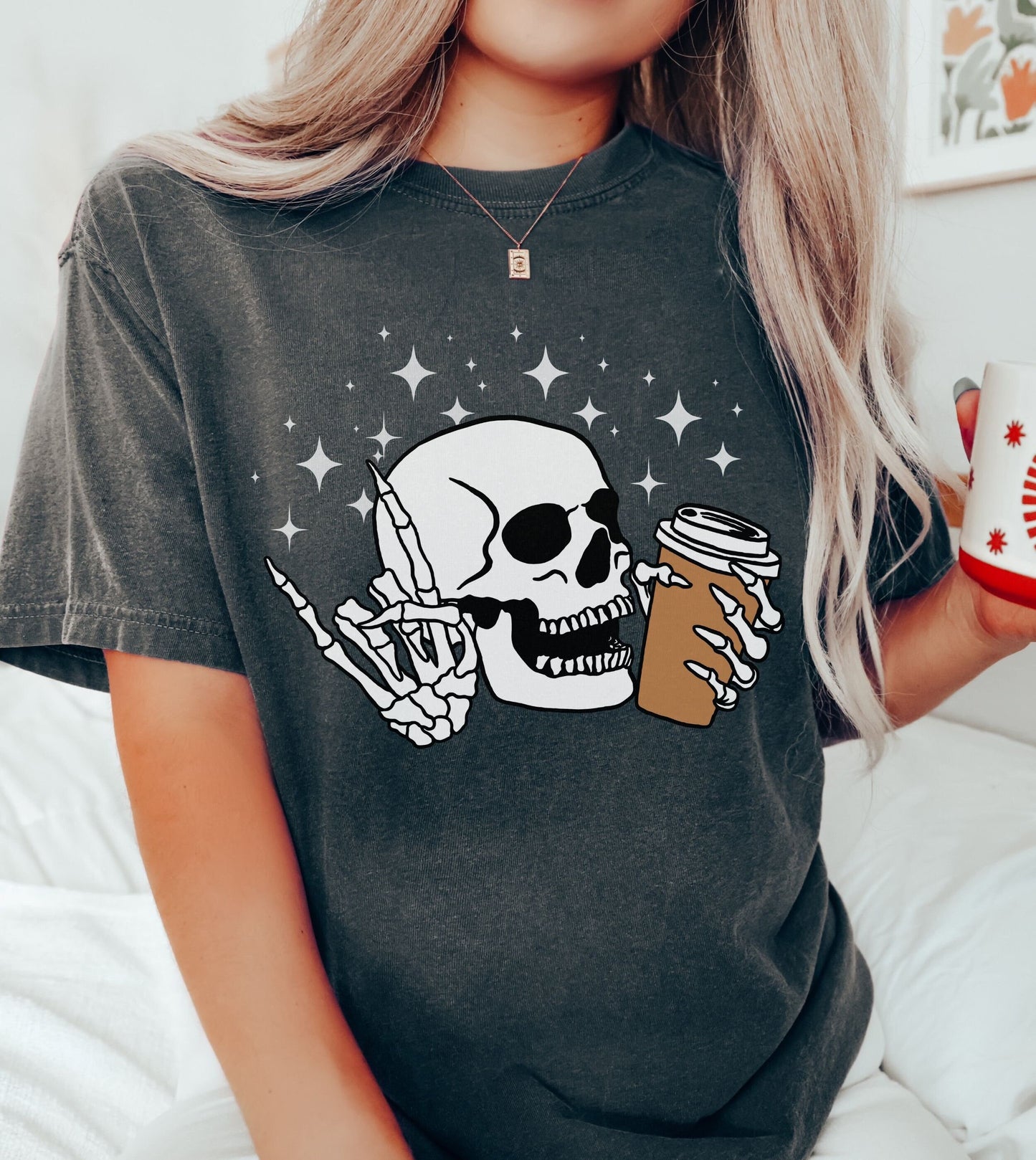 A woman wearing a cute pepper colored shirt, a skeleton skull holding a coffee in the left hand and holding up the rock on hand sign with the right hand with twinkly stars in the background.