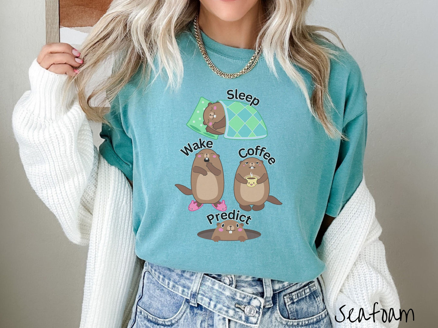 A woman wearing a cute seafoam colored shirt with the text Sleep, Wake, Coffee, and Predict accompanied with pictures of a groundhog sleeping, a groundhog waking up yawning, one holding a steaming cup of coffee, and another exiting its hole.