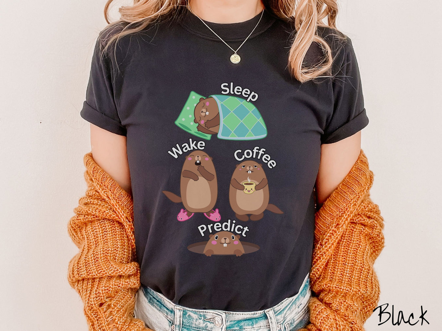 A woman wearing a cute black colored shirt with the text Sleep, Wake, Coffee, and Predict accompanied with pictures of a groundhog sleeping, a groundhog waking up yawning, one holding a steaming cup of coffee, and another exiting its hole.