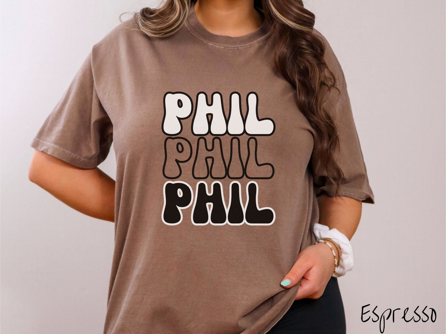 A woman wearing a cute espresso colored shirt with the text PHIL listed three times vertically in white, red, and black color text. The shirt is referring to the chant that occurs for the groundhog Phil every Groundhogs Day in Punxsutawney, PA.