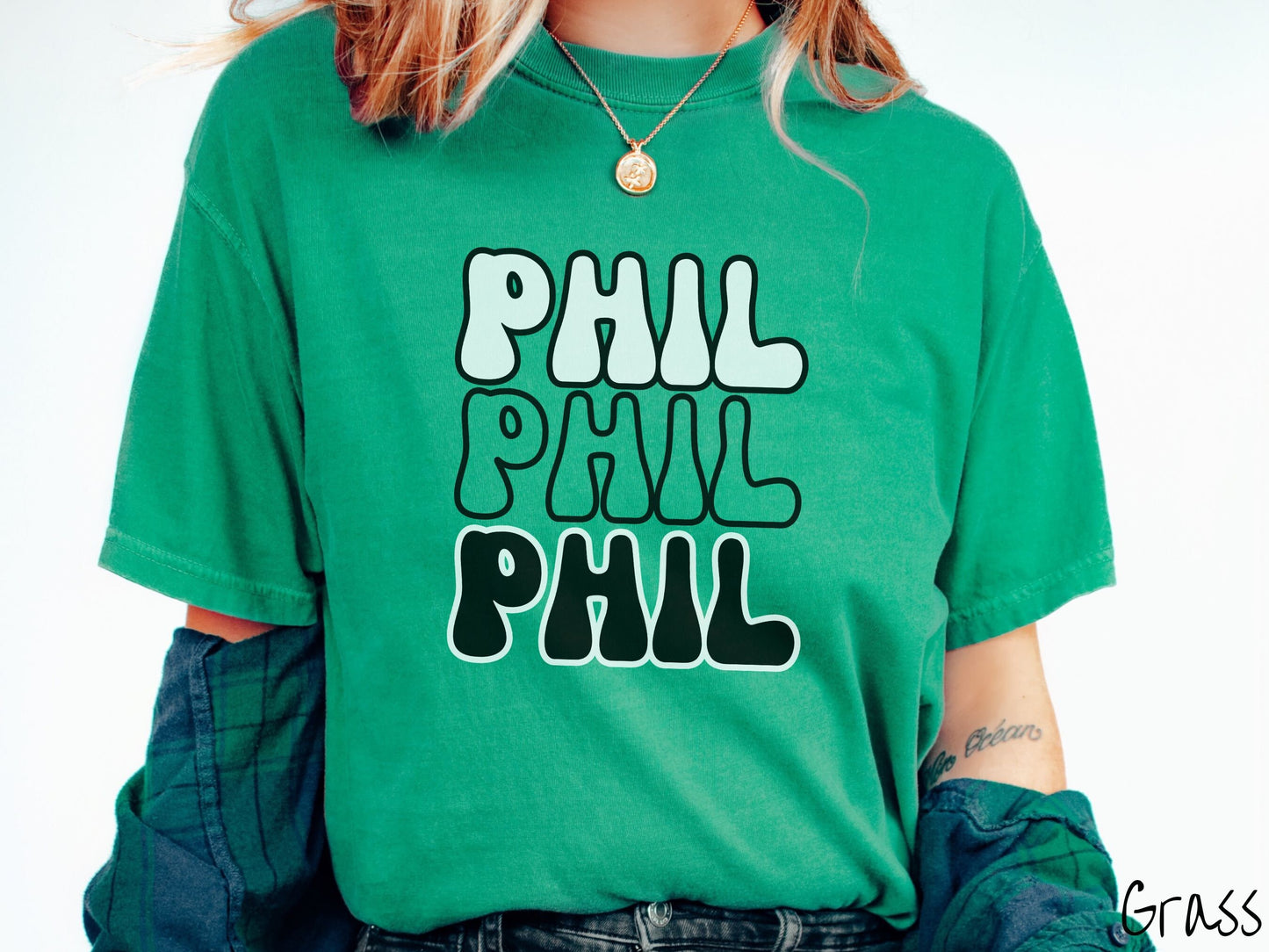A woman wearing a cute grass colored shirt with the text PHIL listed three times vertically in white, red, and black color text. The shirt is referring to the chant that occurs for the groundhog Phil every Groundhogs Day in Punxsutawney, PA.