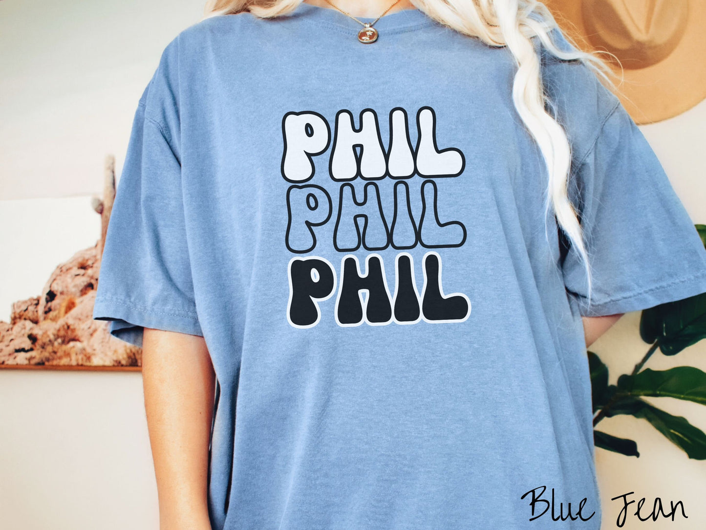 A woman wearing a cute blue jean colored shirt with the text PHIL listed three times vertically in white, red, and black color text. The shirt is referring to the chant that occurs for the groundhog Phil every Groundhogs Day in Punxsutawney, PA.