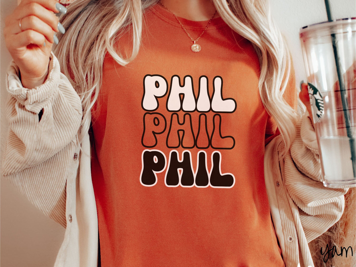 A woman wearing a cute yam colored shirt with the text PHIL listed three times vertically in white, red, and black color text. The shirt is referring to the chant that occurs for the groundhog Phil every Groundhogs Day in Punxsutawney, PA.