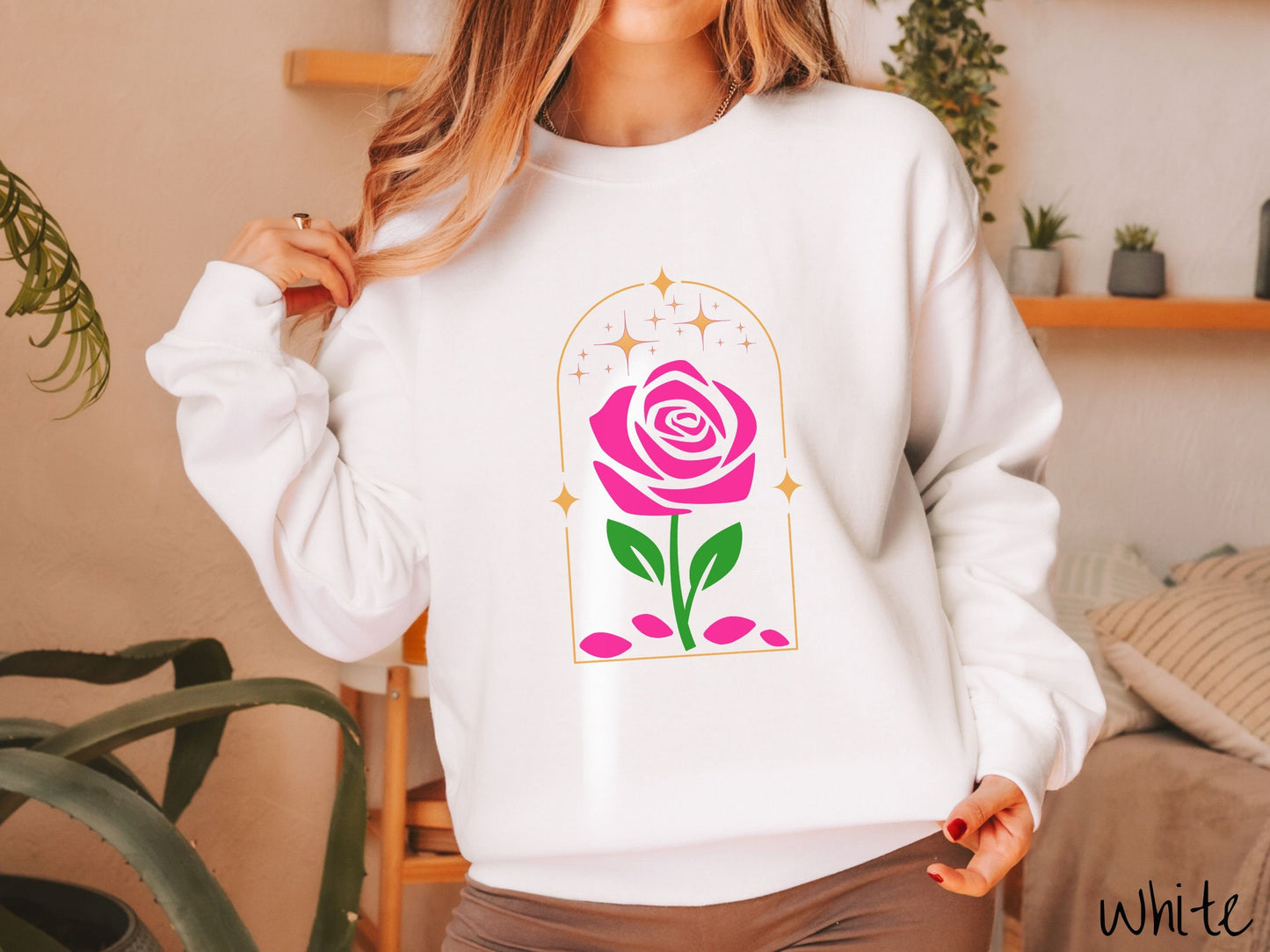 A woman wearing a cute, vintage white colored sweatshirt with an enchanted pink rose in a golden clear container with gold stars twinkling above and pink flower petals resting below.