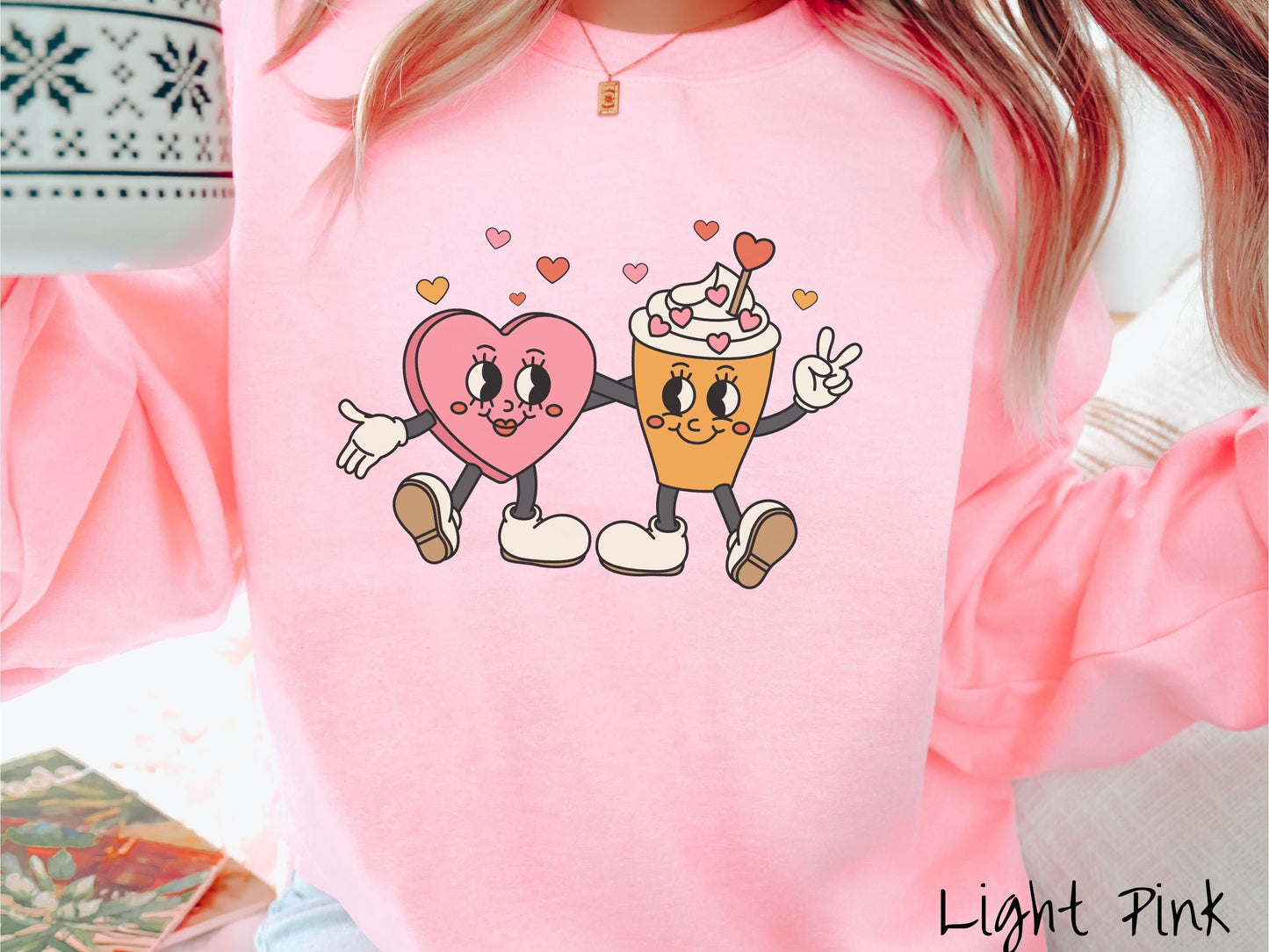A woman wearing a cute, vintage light pink colored sweatshirt with colorful hearts above, a female heart with smiling face, legs, and arms wrapping her left arm around a whipped cream topped light brown coffee cup with a heart-tipped topper.