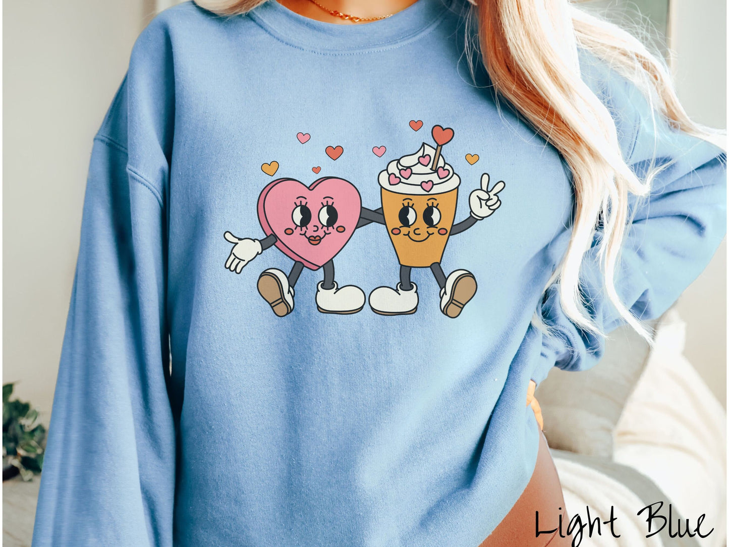 A woman wearing a cute, vintage light blue colored sweatshirt with colorful hearts above, a female heart with smiling face, legs, and arms wrapping her left arm around a whipped cream topped light brown coffee cup with a heart-tipped topper.