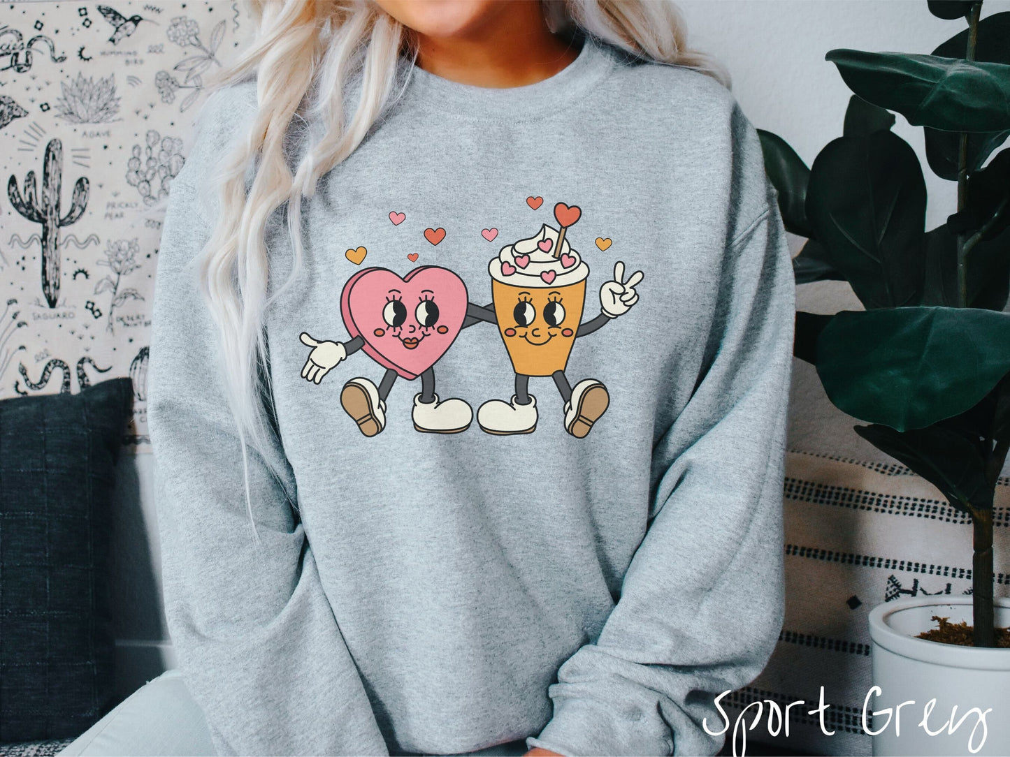 A woman wearing a cute, vintage sport grey colored sweatshirt with colorful hearts above, a female heart with smiling face, legs, and arms wrapping her left arm around a whipped cream topped light brown coffee cup with a heart-tipped topper.