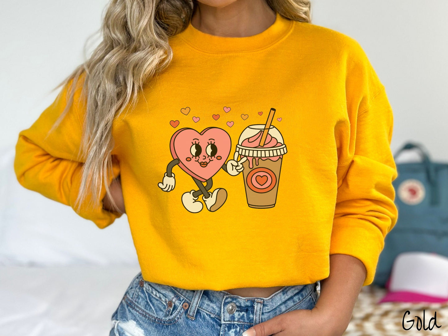 A woman wearing a cute, vintage gold colored sweatshirt and colorful hearts above, a female heart with smiling face, legs, and arms showing a peace sign with her left hand next to an iced coffee and pink whipped cream and a red heart logo on the cup.