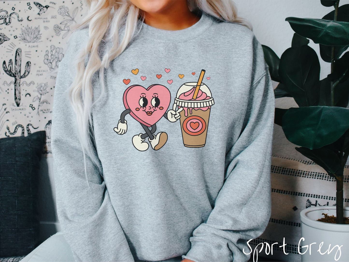 A woman wearing a cute, vintage sport grey colored sweatshirt and hearts above, a female heart with smiling face, legs, and arms showing a peace sign with her left hand next to an iced coffee and pink whipped cream and a red heart logo on the cup.