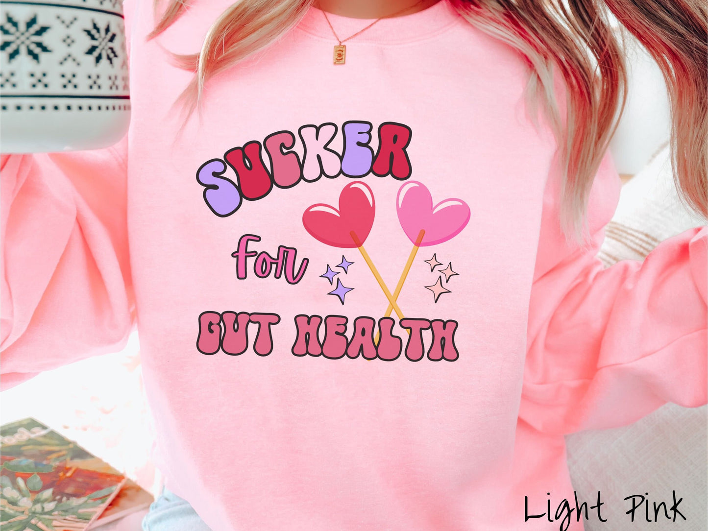 A woman wearing a cute, vintage light pink colored sweatshirt with the text Sucker for Gut Health in red, pink, and purple, and there are two lollipops with pink and red hearts on the ends of the sticks and purple and yellow stars in the background.