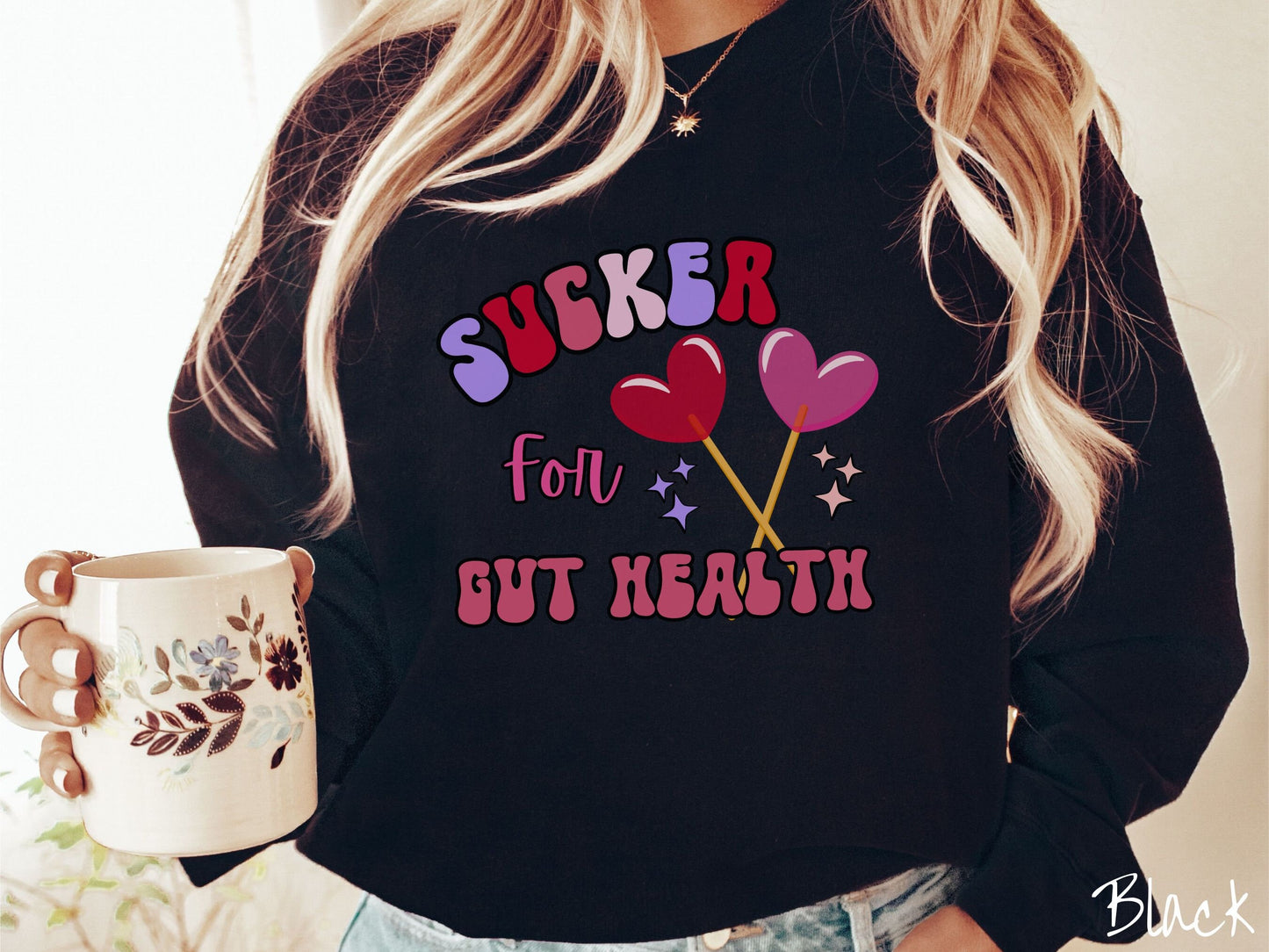 A woman wearing a cute, vintage black colored sweatshirt with the text Sucker for Gut Health in red, pink, and purple, and there are two lollipops with pink and red hearts on the ends of the sticks and purple and yellow stars in the background.