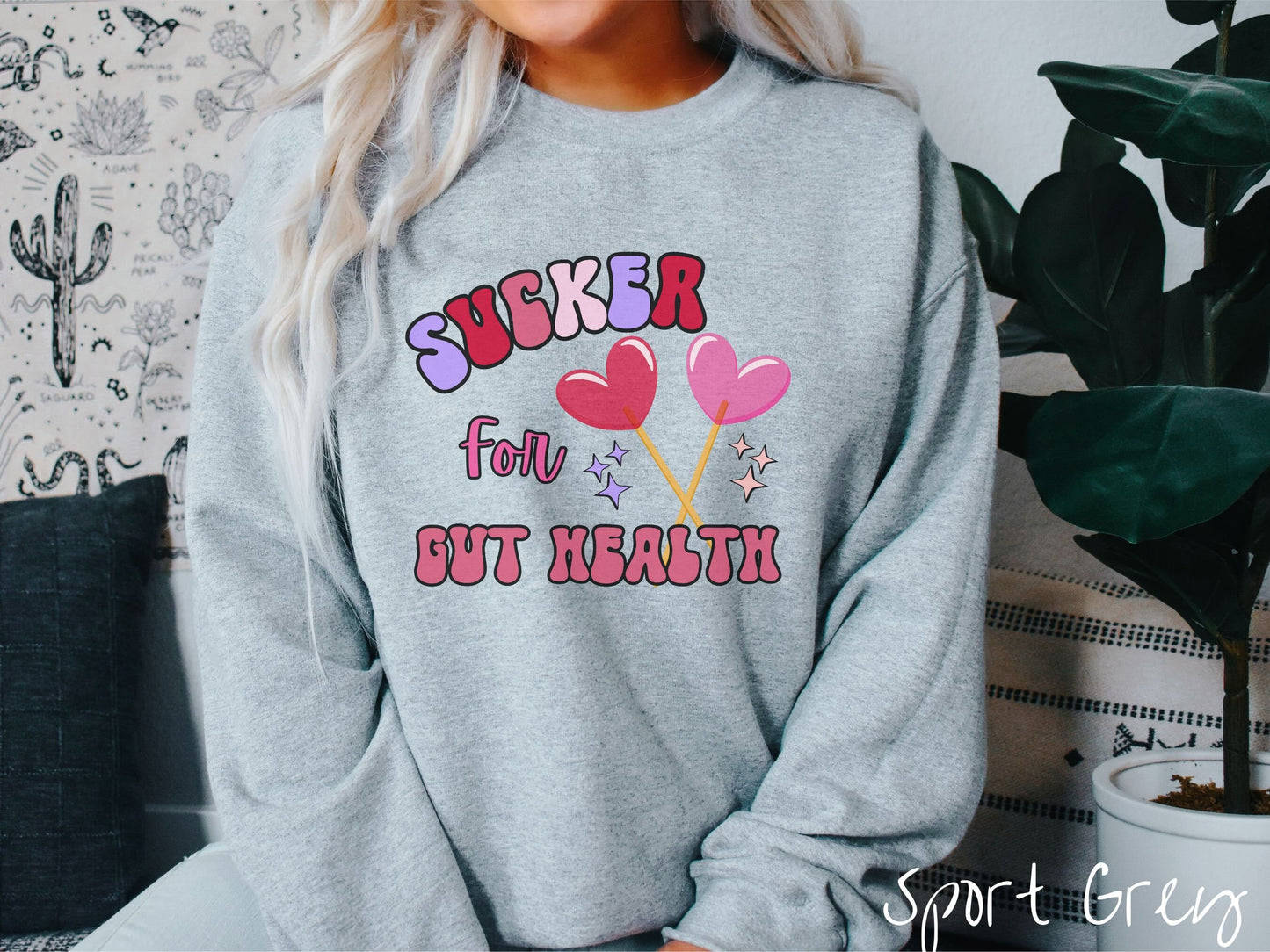 A woman wearing a cute, vintage sport grey colored sweatshirt with the text Sucker for Gut Health in red, pink, and purple, and there are two lollipops with pink and red hearts on the ends of the sticks and purple and yellow stars in the background.