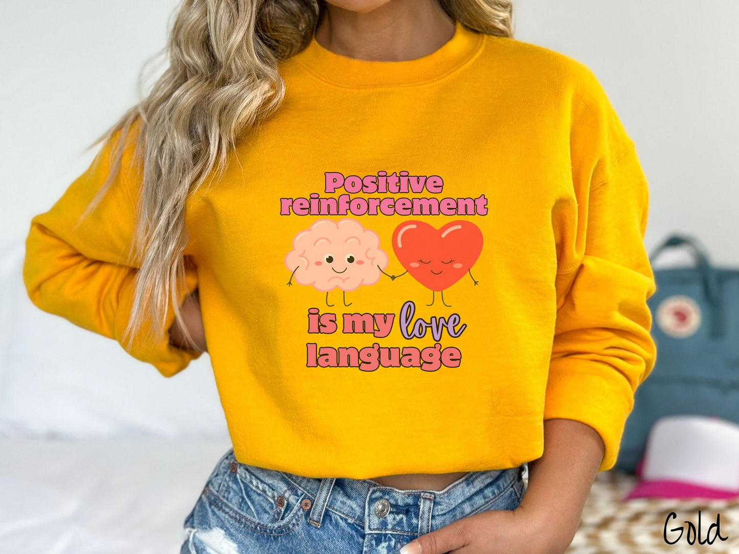 A woman wearing a cute, vintage gold colored sweatshirt with the text Positive reinforcement is my love language in pink, purple, and red with a pink smiling brain holding hands with a red smiling heart.