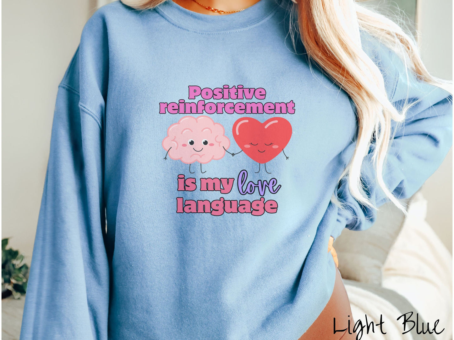 A woman wearing a cute, vintage light blue colored sweatshirt with the text Positive reinforcement is my love language in pink, purple, and red with a pink smiling brain holding hands with a red smiling heart.
