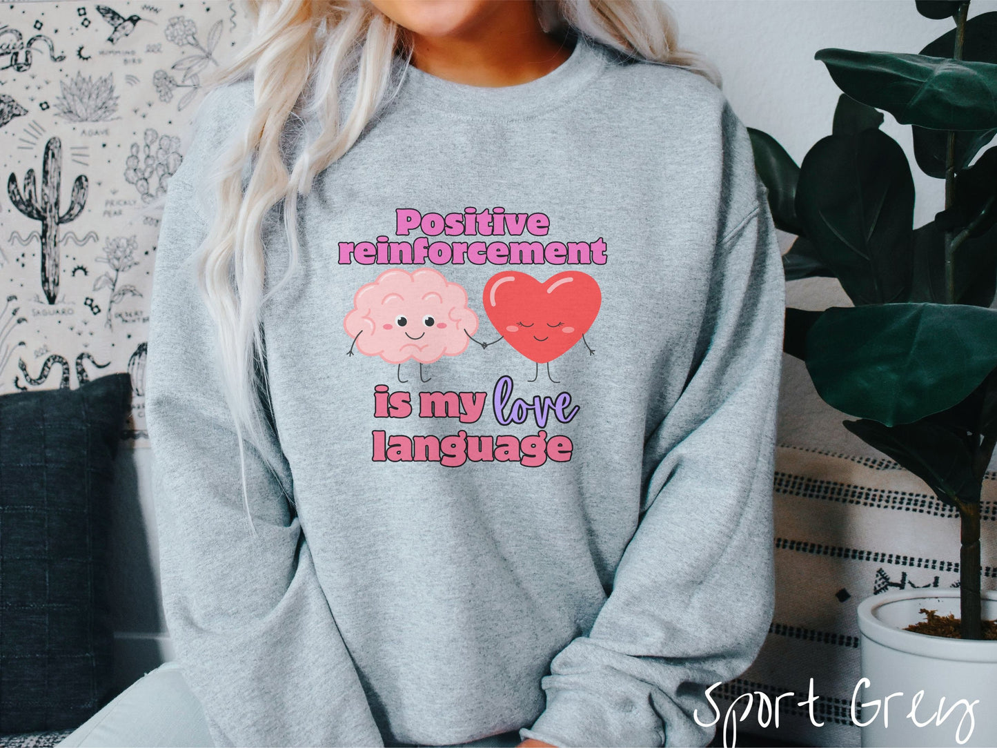 A woman wearing a cute, vintage sport grey colored sweatshirt with the text Positive reinforcement is my love language in pink, purple, and red with a pink smiling brain holding hands with a red smiling heart.