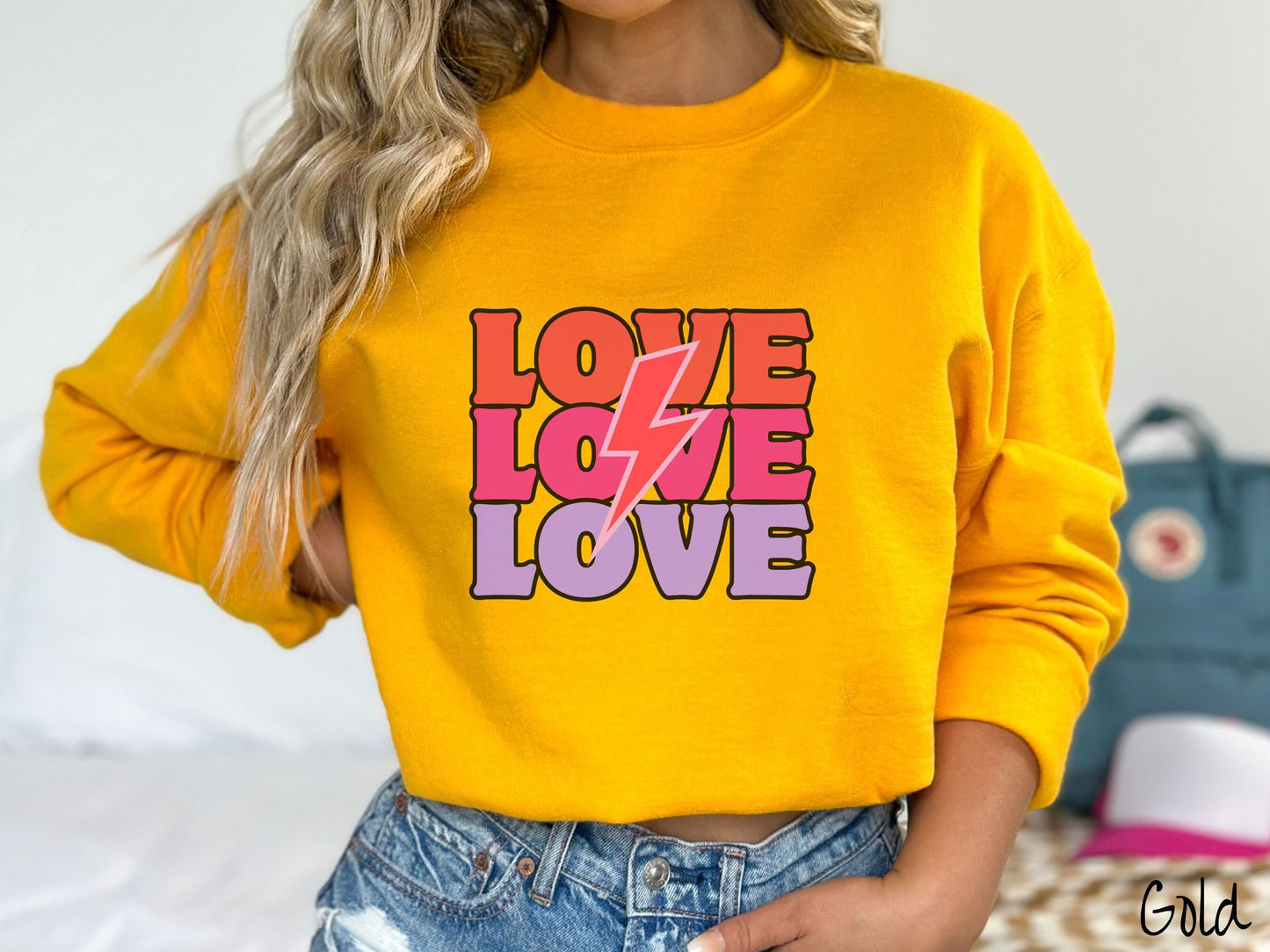 A woman wearing a cute, vintage gold colored sweatshirt with the word Love listed three times vertically with a pink lightning bolt going through the middle of the words.