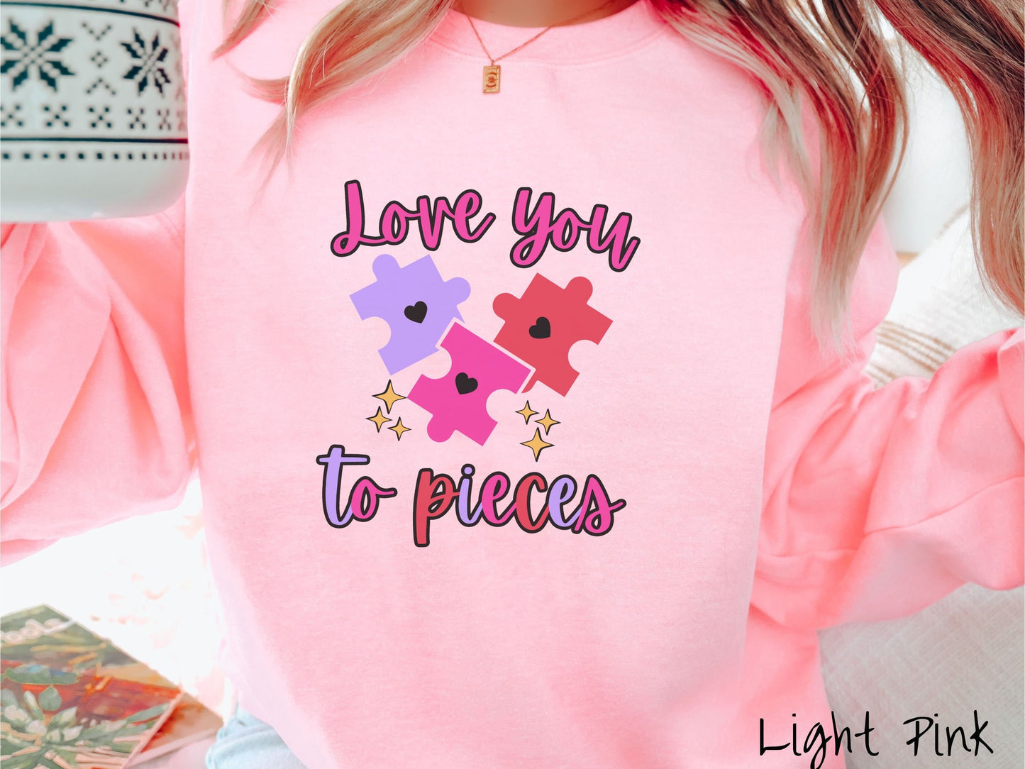 A woman wearing a cute, vintage light pink colored sweatshirt with the text Love you to pieces in red, pink, and purple colors, and three red, pink, and purple puzzle pieces with hearts on them surrounded by yellow stars.