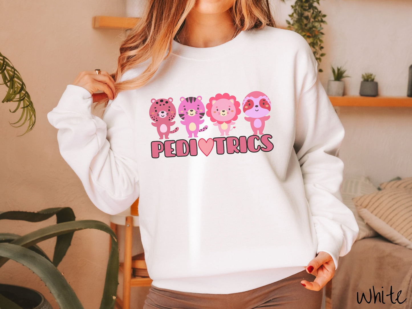 A woman wearing a cute white colored sweatshirt with the text Pediatrics in the center, a heart replacing the letter A, and above the text are a maroon lion, a pink tiger, a red lion, and a pink sloth all with their hands open as if asking for a hug.