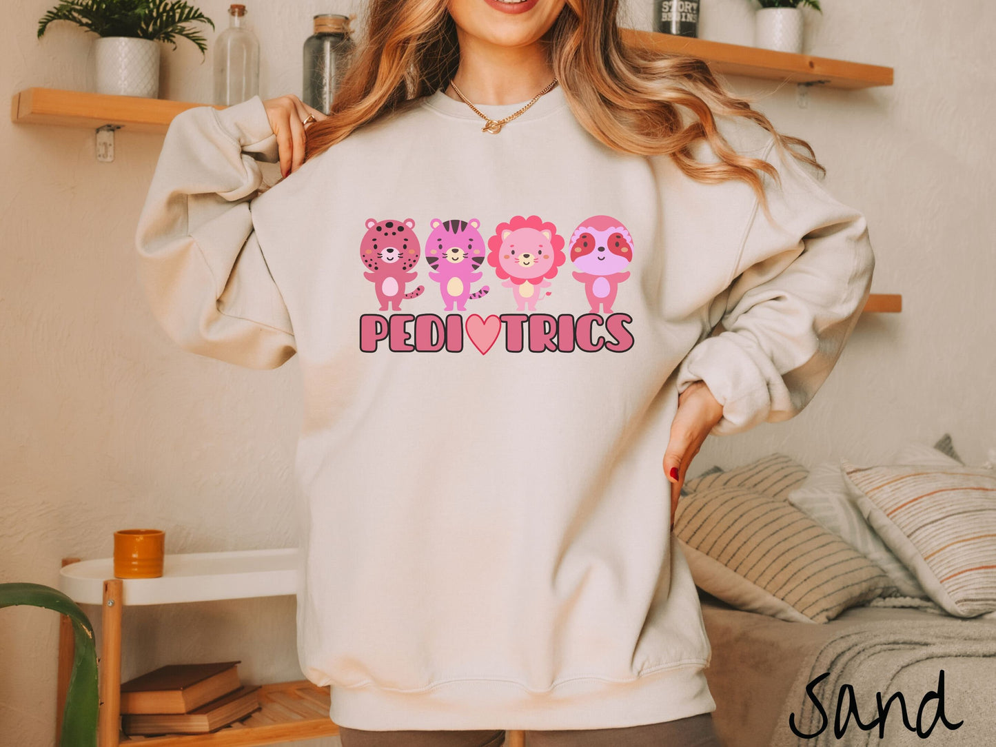 A woman wearing a cute sand colored sweatshirt with the text Pediatrics in the center, a heart replacing the letter A, and above the text are a maroon lion, a pink tiger, a red lion, and a pink sloth all with their hands open asking for a hug.