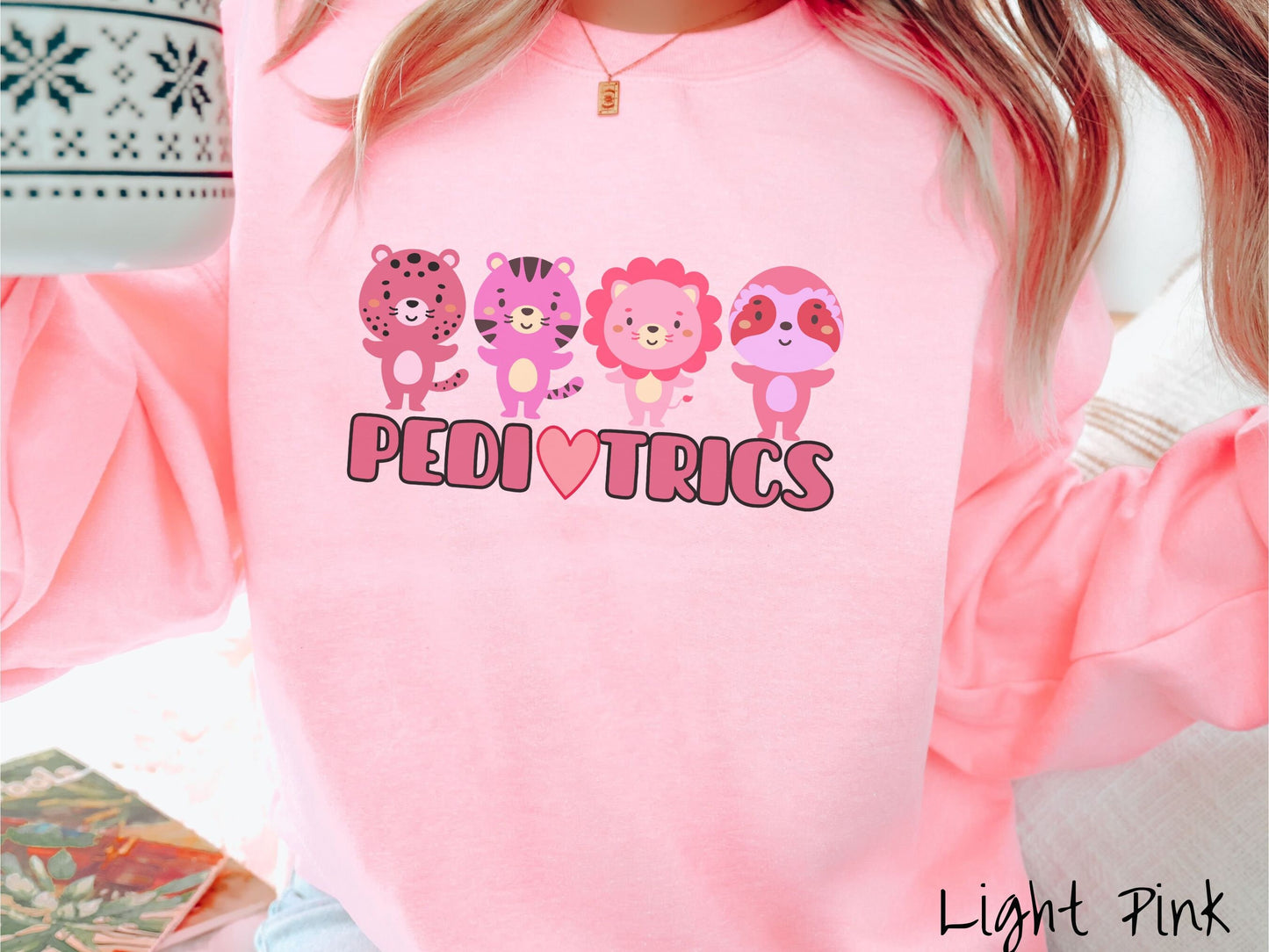 A woman wearing a cute light pink colored sweatshirt with the text Pediatrics in the center, a heart replacing the letter A, and above the text are a maroon lion, a pink tiger, a red lion, and a pink sloth all with their hands open asking for a hug.