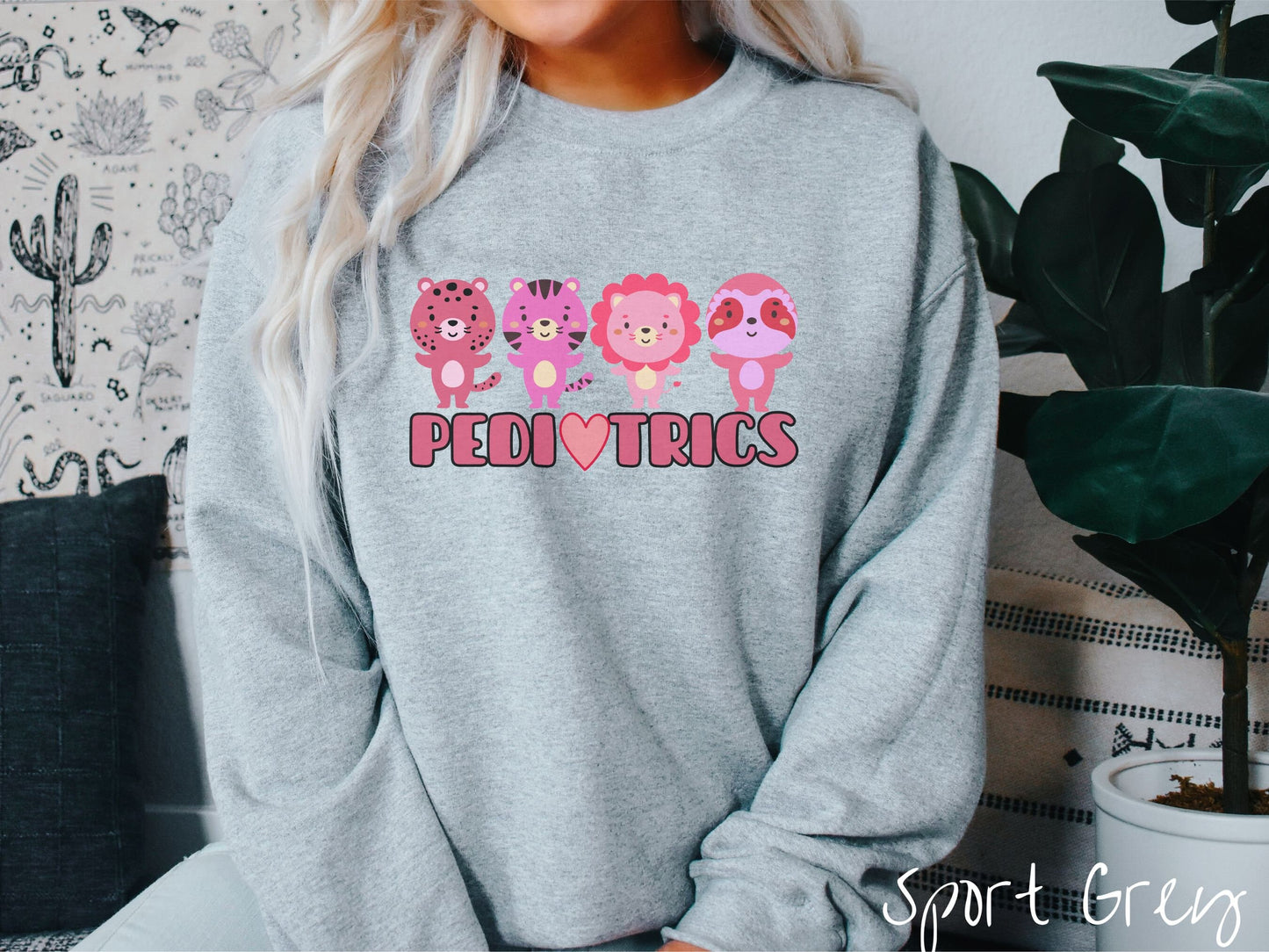 A woman wearing a cute sport grey colored sweatshirt with the text Pediatrics in the center, a heart replacing the letter A, and above the text are a maroon lion, a pink tiger, a red lion, and a pink sloth all with their hands open asking for a hug.