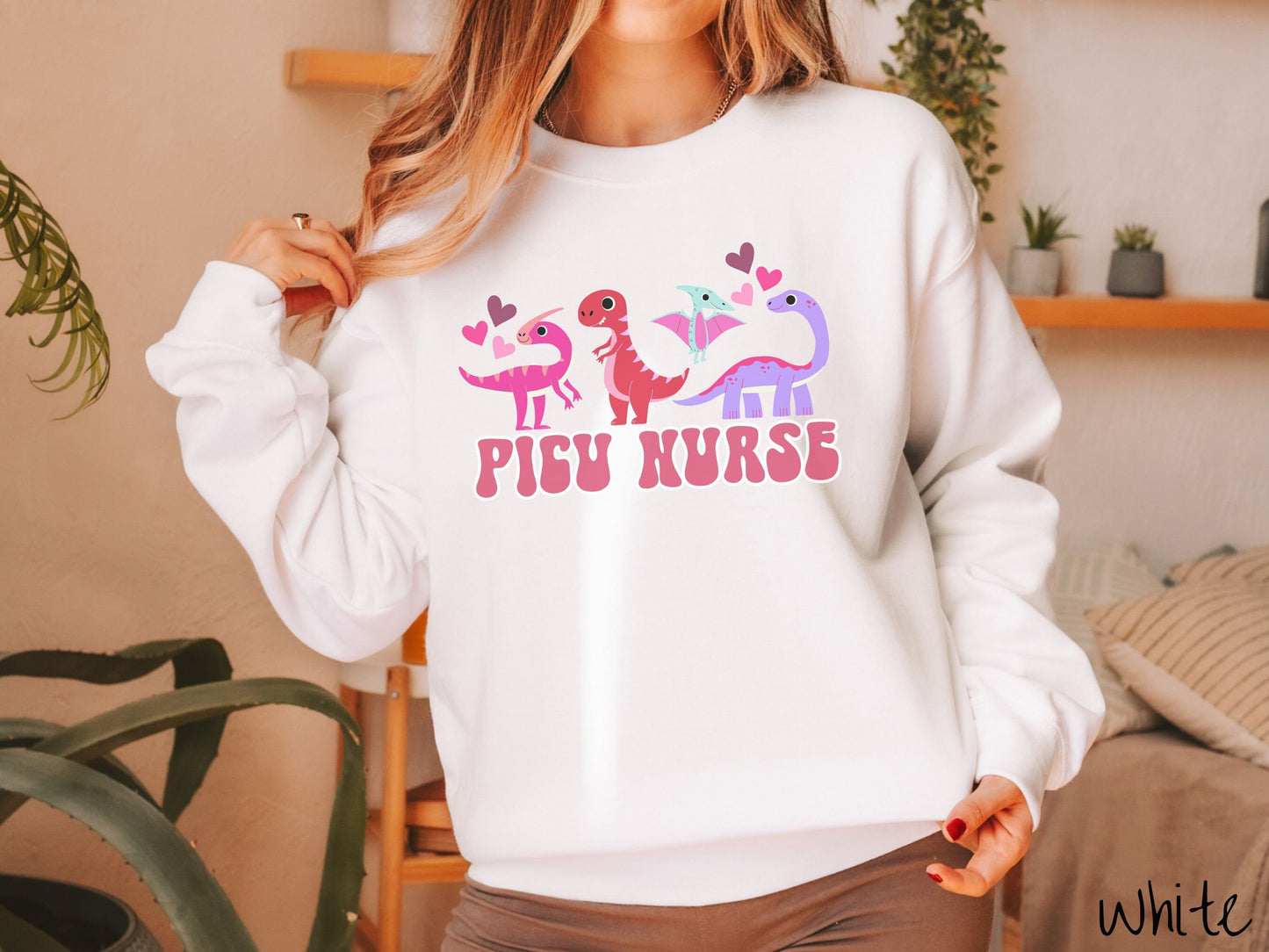 A woman wearing a cute white colored sweatshirt with the text PICU Nurse in the center below four dinosaurs, a pink Parasaurolophus, a red tyrannosaurus rex, a green pterodactyl, a purple Brachiosaurus, and pink and red hearts in the air.
