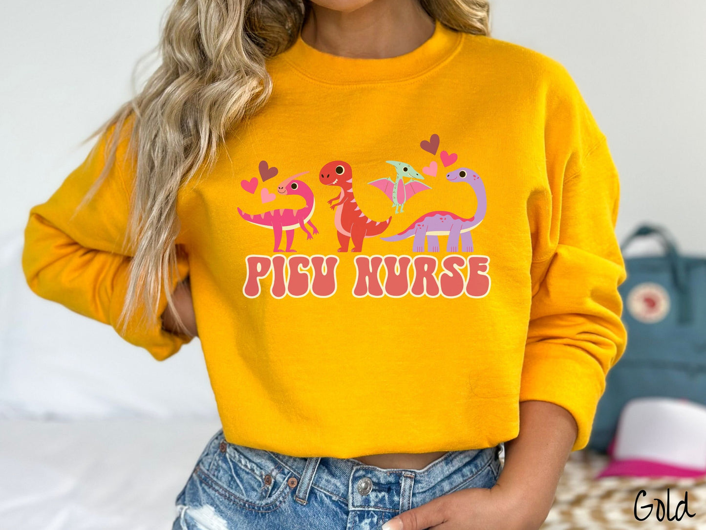 A woman wearing a cute gold colored sweatshirt with the text PICU Nurse in the center below four dinosaurs, a pink Parasaurolophus, a red tyrannosaurus rex, a green pterodactyl, a purple Brachiosaurus, and pink and red hearts in the air.