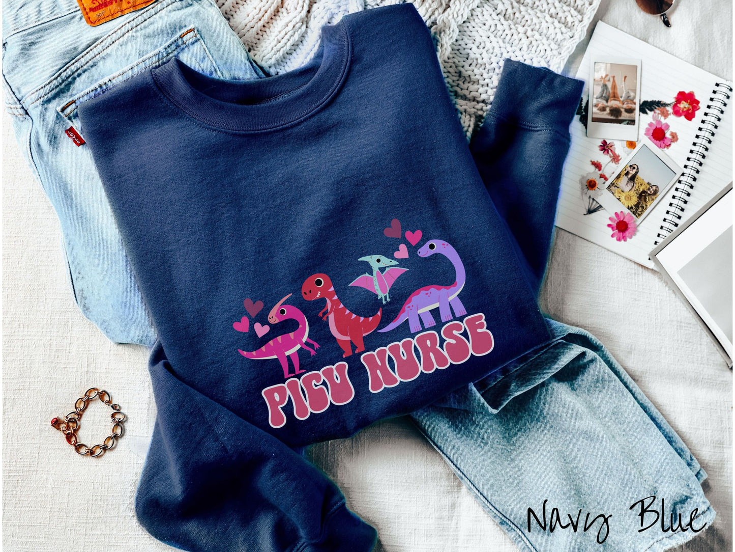 A navy blue colored sweatshirt with the text PICU Nurse in the center below four dinosaurs, a pink Parasaurolophus, a red tyrannosaurus rex, a green pterodactyl, a purple Brachiosaurus, and pink and red hearts in the air.