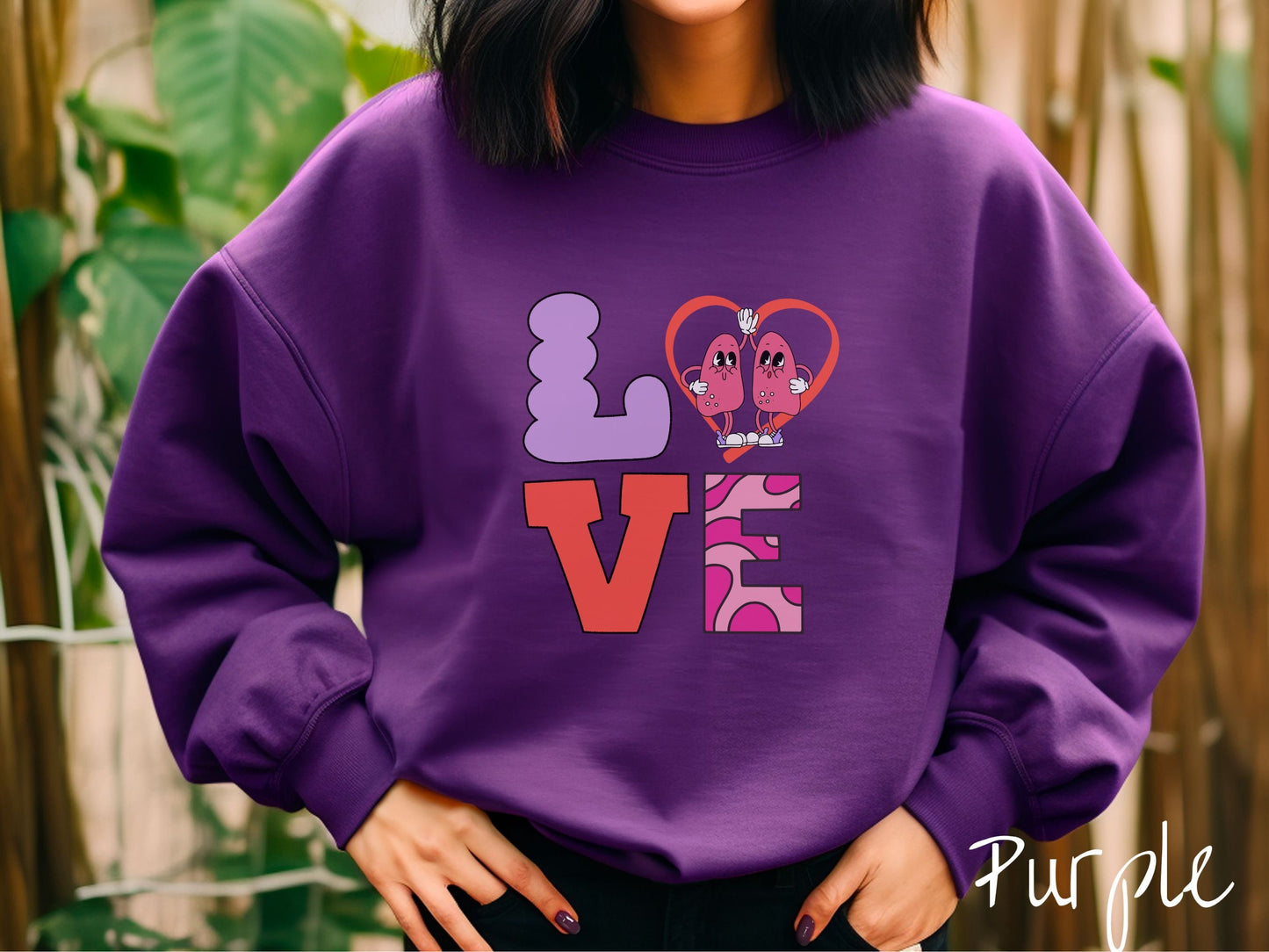 A woman wearing a cute purple colored sweatshirt with the word LOVE spelled out with the L as a purple lung, the O as a heart with two lung figures inside high fiving, the V is orange, and the E is two shades of pink.