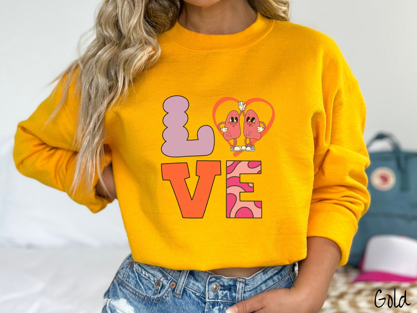 A woman wearing a cute gold colored sweatshirt with the word LOVE spelled out with the L as a purple lung, the O as a heart with two lung figures inside high fiving, the V is orange, and the E is two shades of pink.