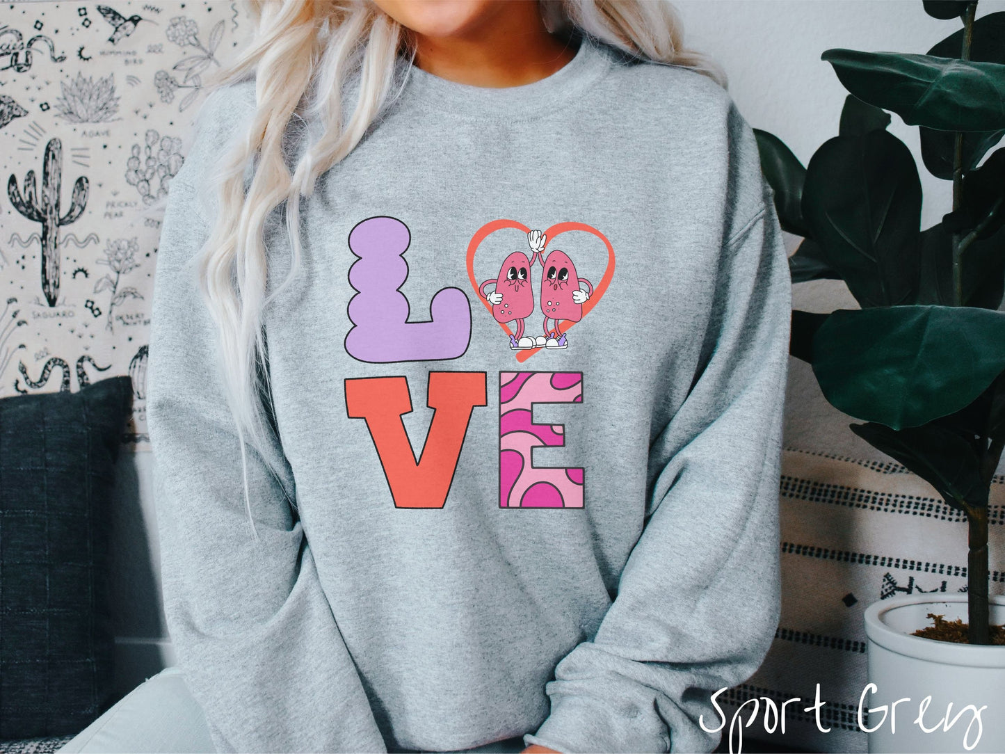 A woman wearing a cute sport grey colored sweatshirt with the word LOVE spelled out with the L as a purple lung, the O as a heart with two lung figures inside high fiving, the V is orange, and the E is two shades of pink.
