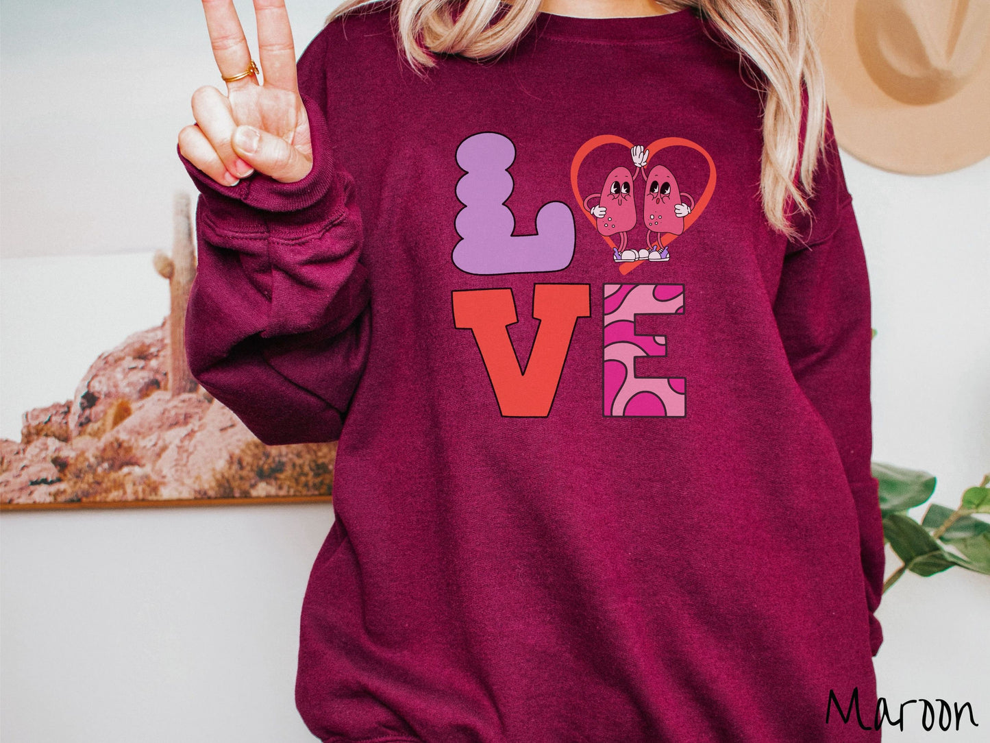 A woman wearing a cute maroon colored sweatshirt with the word LOVE spelled out with the L as a purple lung, the O as a heart with two lung figures inside high fiving, the V is orange, and the E is two shades of pink.