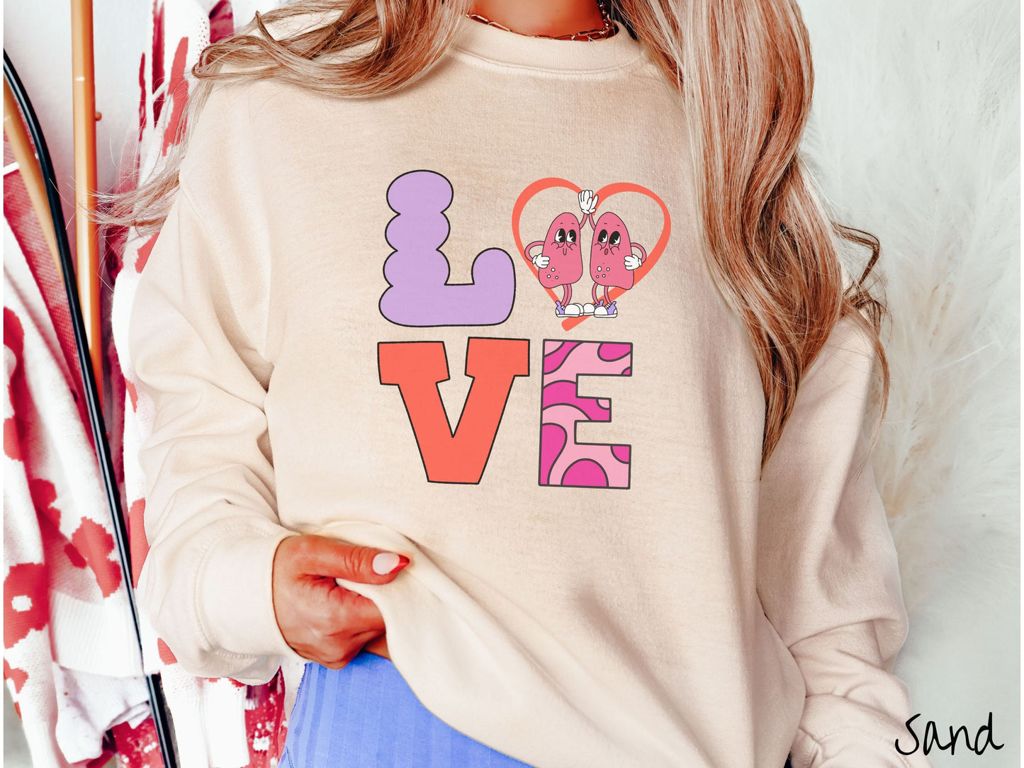 A woman wearing a cute sand colored sweatshirt with the word LOVE spelled out with the L as a purple lung, the O as a heart with two lung figures inside high fiving, the V is orange, and the E is two shades of pink.