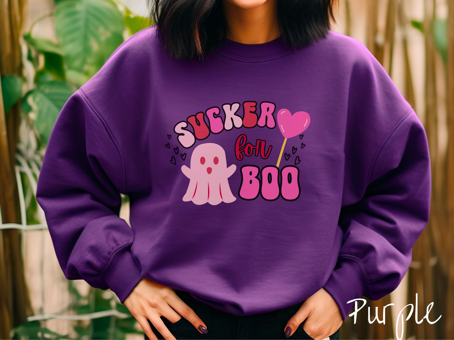 A woman wearing a cute purple colored sweatshirt with the text saying Sucker for Boo, with a pink ghost with its arms open, a lollipop with a pink heart, and hearts floating in the air.