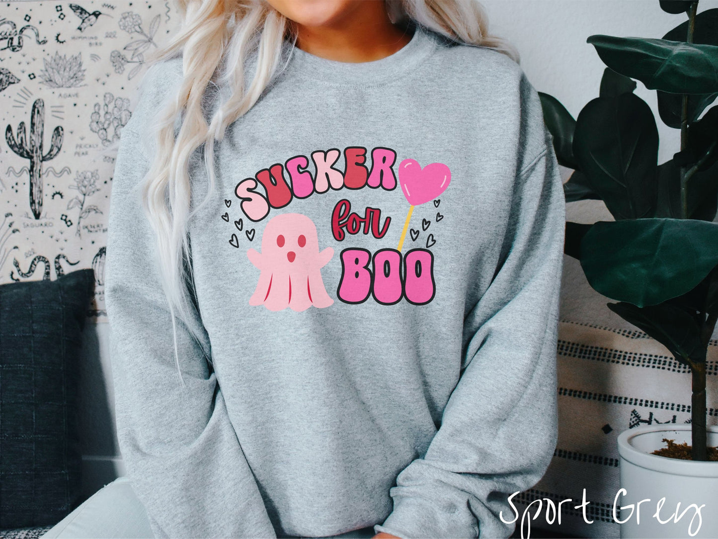 A woman wearing a cute sport grey colored sweatshirt with the text saying Sucker for Boo, with a pink ghost with its arms open, a lollipop with a pink heart, and hearts floating in the air.
