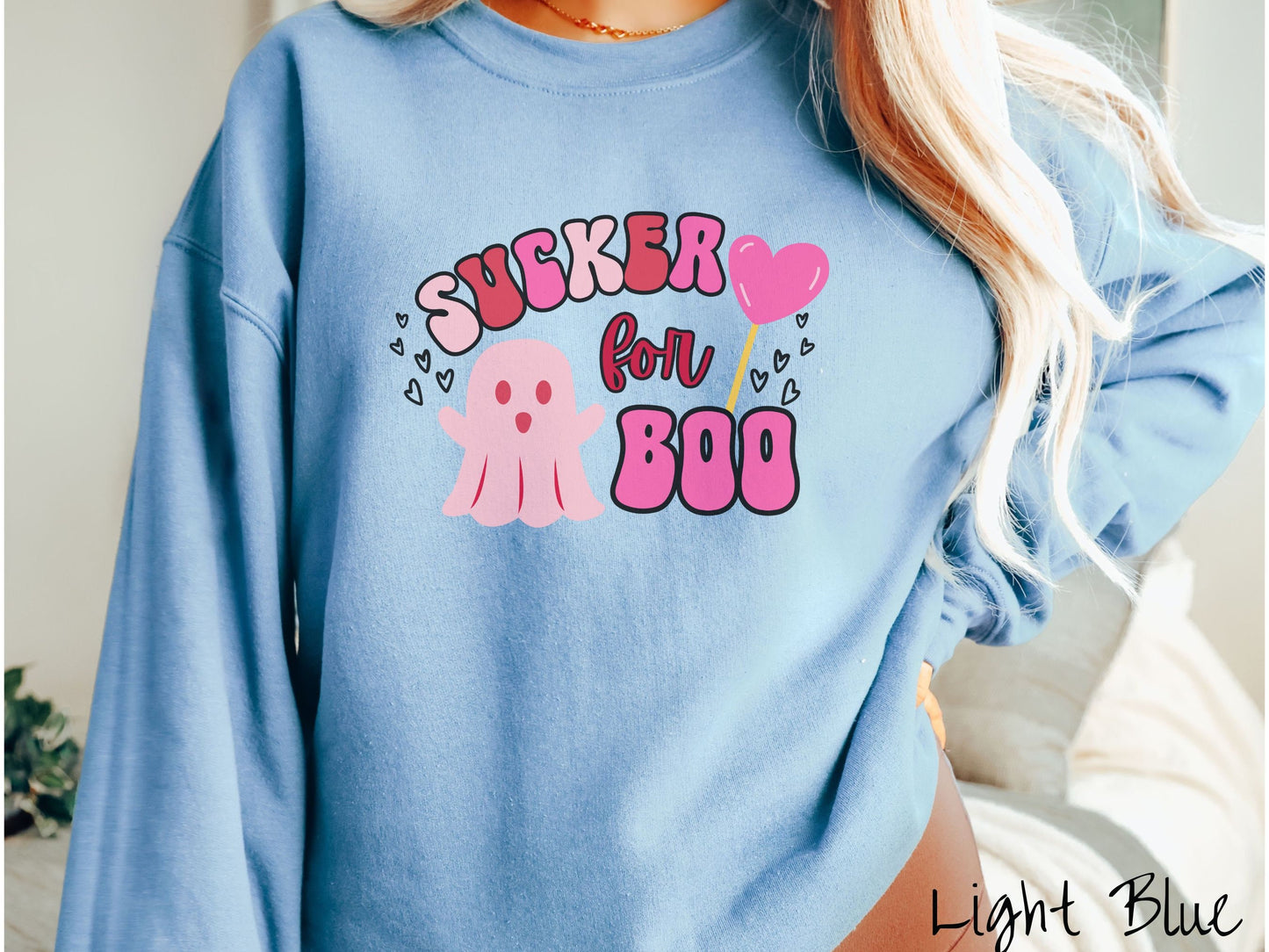 A woman wearing a cute light blue colored sweatshirt with the text saying Sucker for Boo, with a pink ghost with its arms open, a lollipop with a pink heart, and hearts floating in the air.